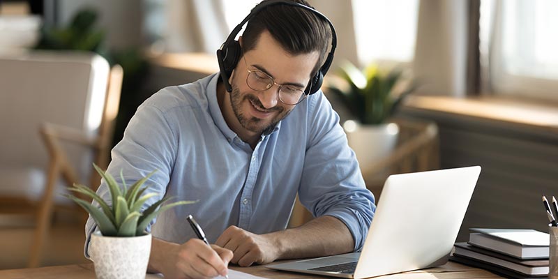 Male employee sitting at desk with headphones, learning on the computer and taking notes.