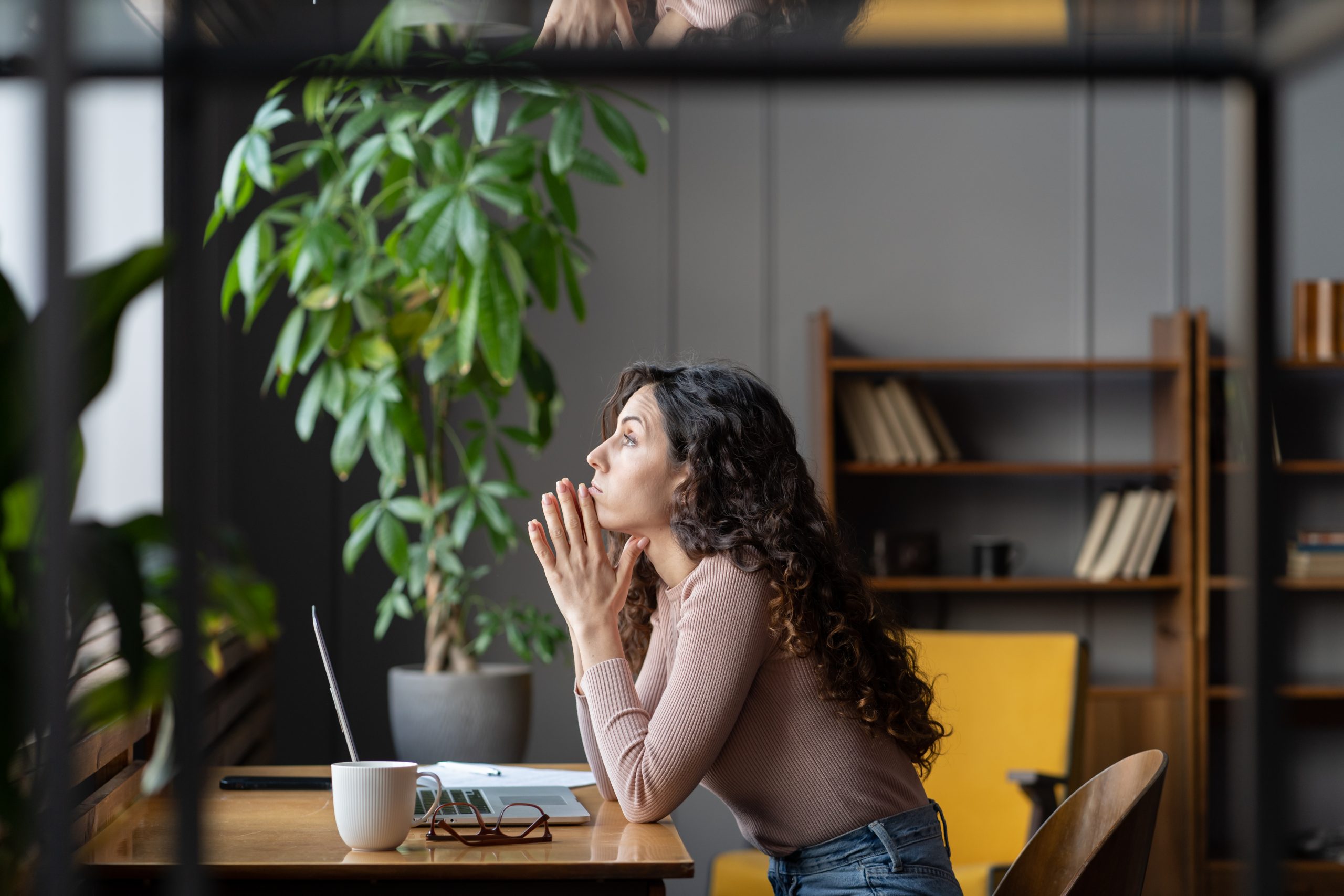 A female employee sits at her desk but is disengaged in work and is looking out the window.