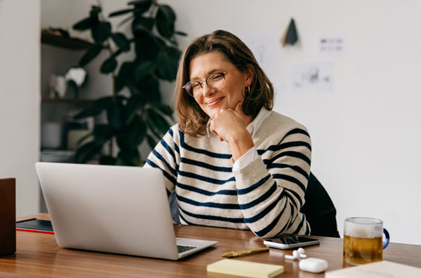A middle-aged woman uses technology at her desk with laptop and notes nearby, capturing productivity, communication, and business focus in a cozy home office.